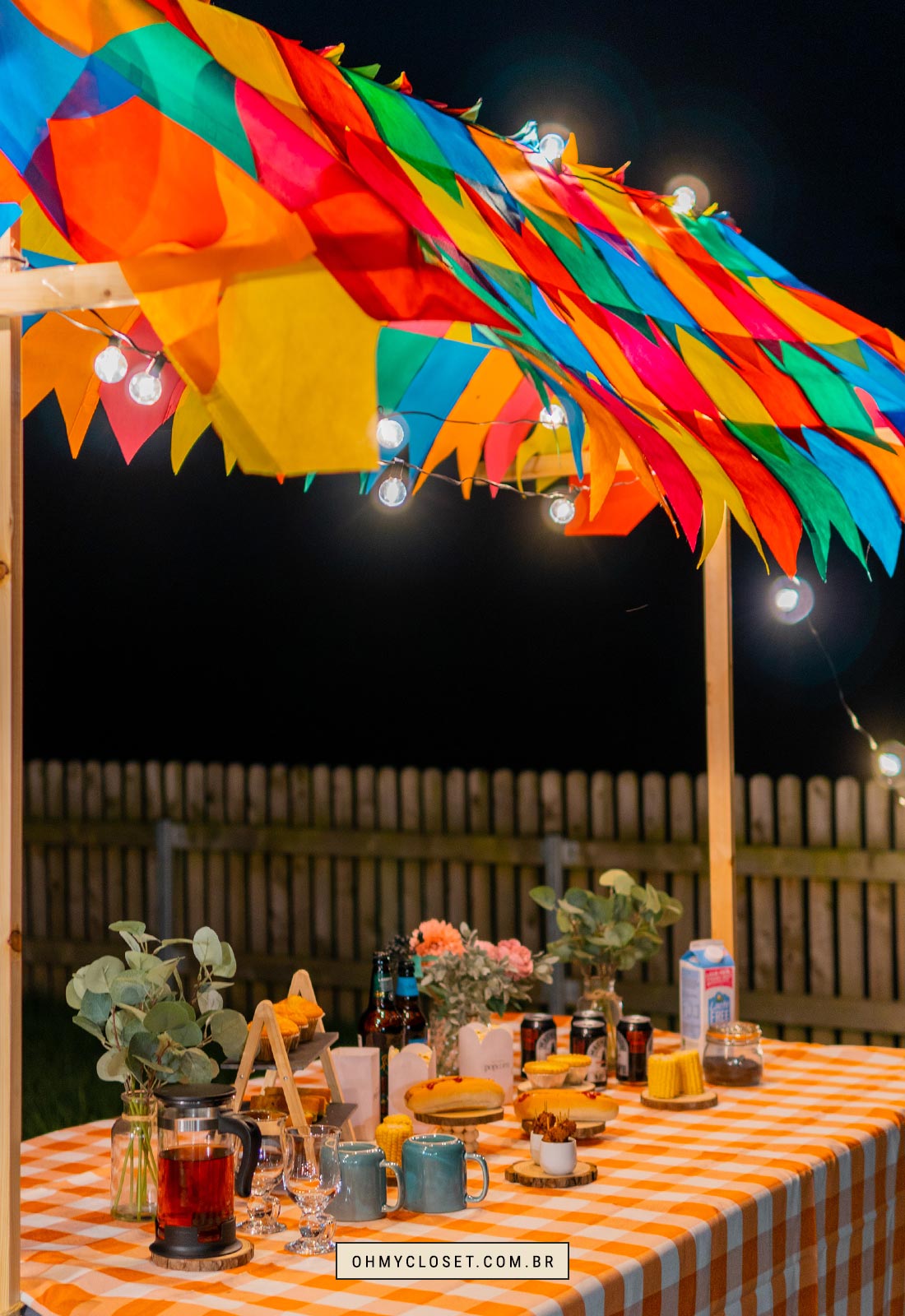 Mesa de festa junina decorada com comidas e bebidas com varal de lâmpadas.